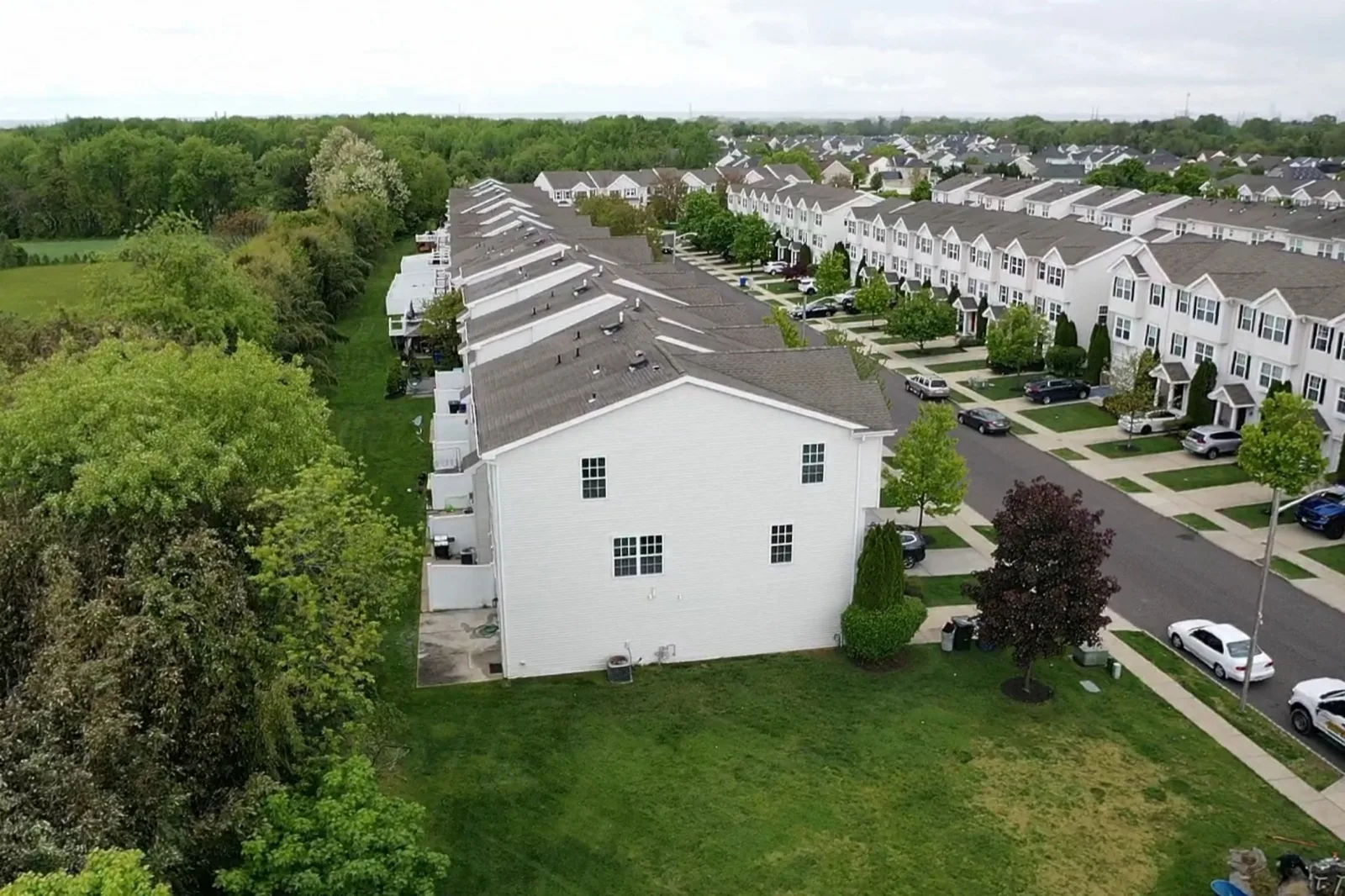 An aerial of rows of attached townhomes along a curving street, with lawns and woods beside them